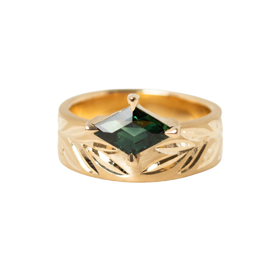A 14k gold ring featuring a hexagon dark green sapphire in a prong eagle setting, with an hand engraved leaf pattern on the band, shown against a plain white background.