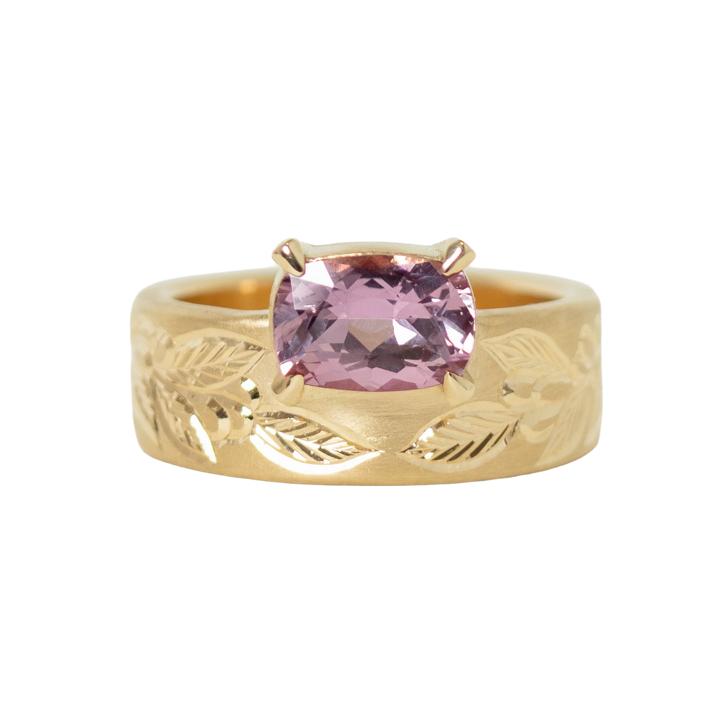 A 14k yellow gold ring featuring a cushion-cut pink sapphire in a prong setting, with an hand engraved leaf pattern on the band, shown against a plain white background.