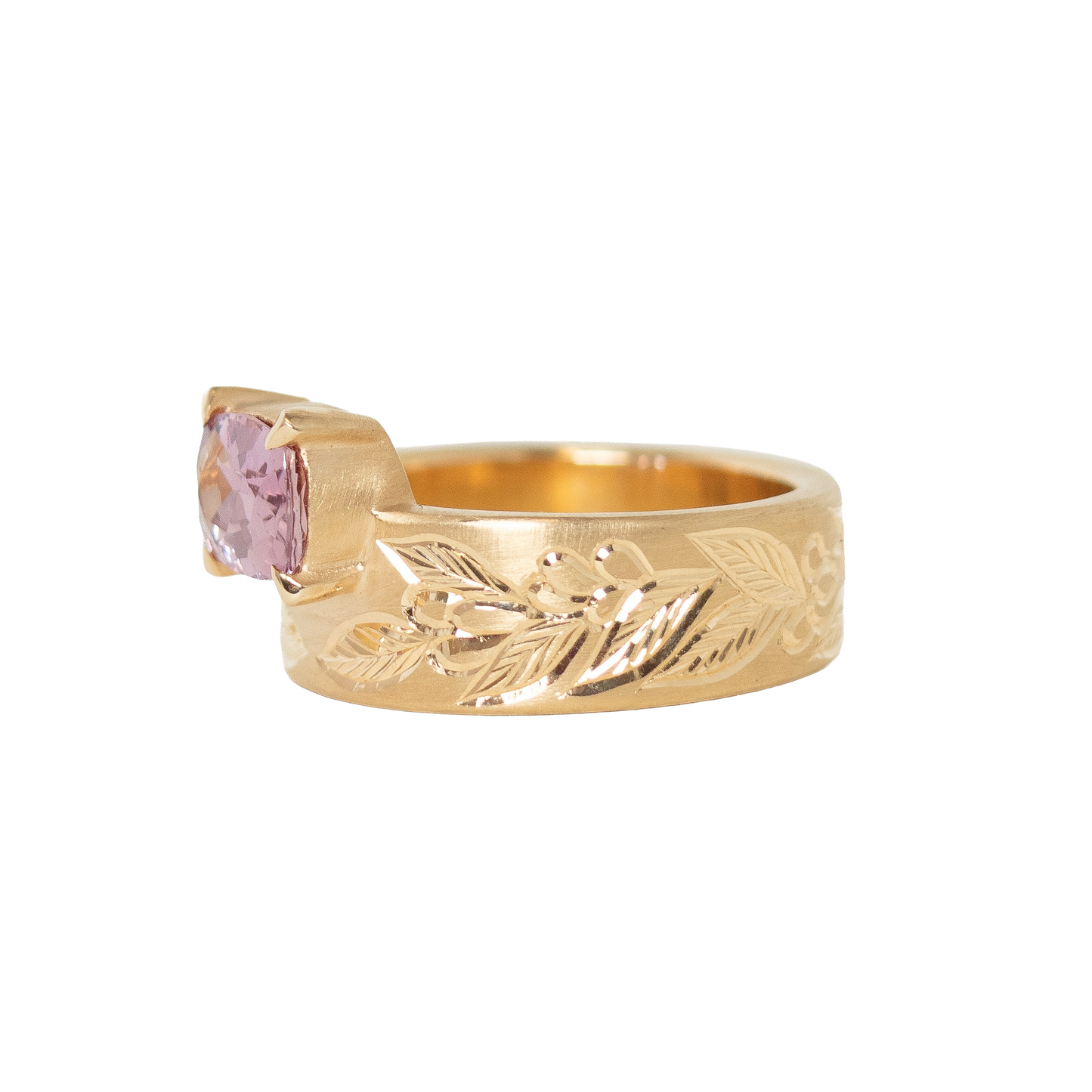 A 14k yellow gold ring featuring a cushion-cut pink sapphire in a prong setting, with an hand engraved leaf pattern on the band, shown against a plain white background.