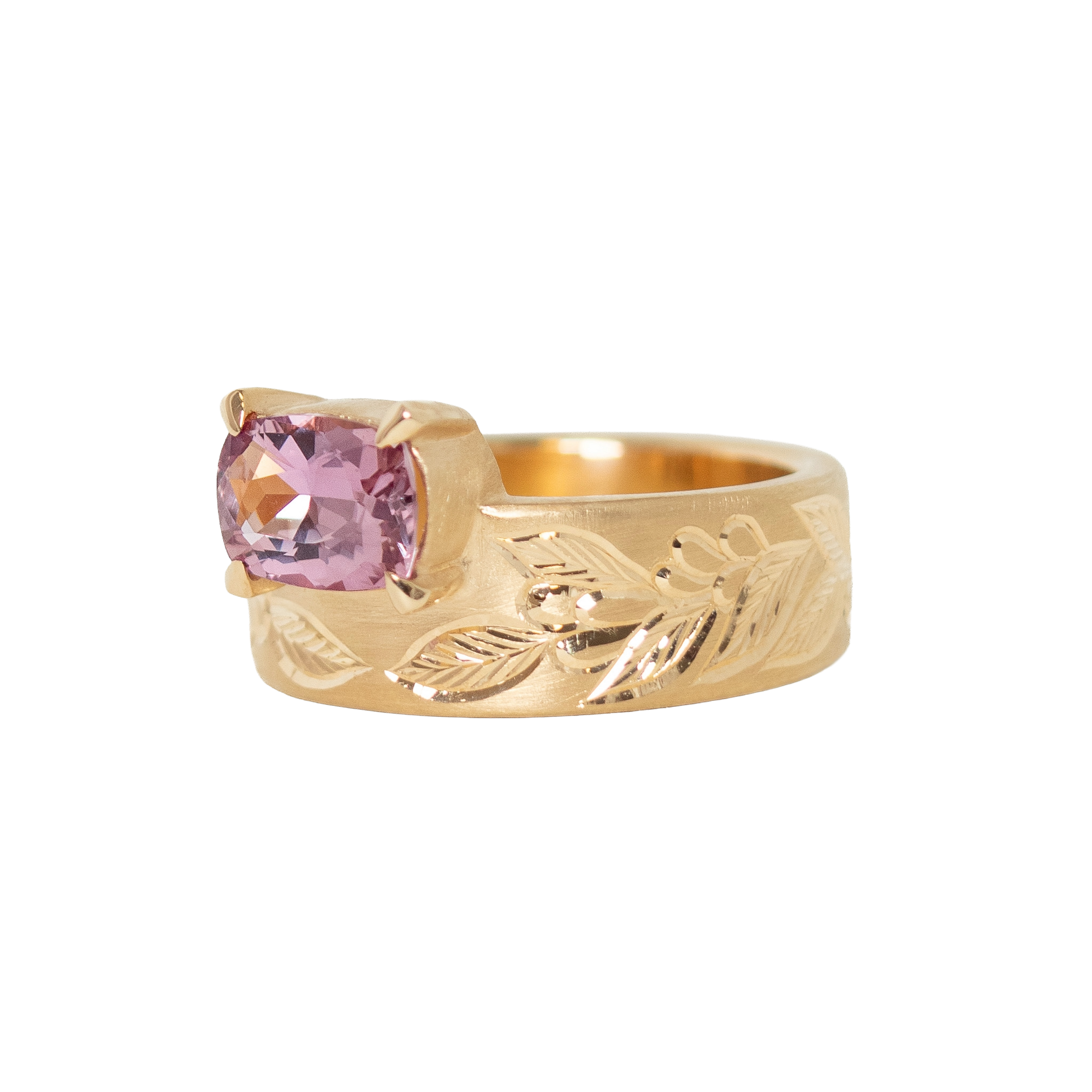 A 14k yellow gold ring featuring a cushion-cut pink sapphire in a prong setting, with an hand engraved leaf pattern on the band, shown against a plain white background.