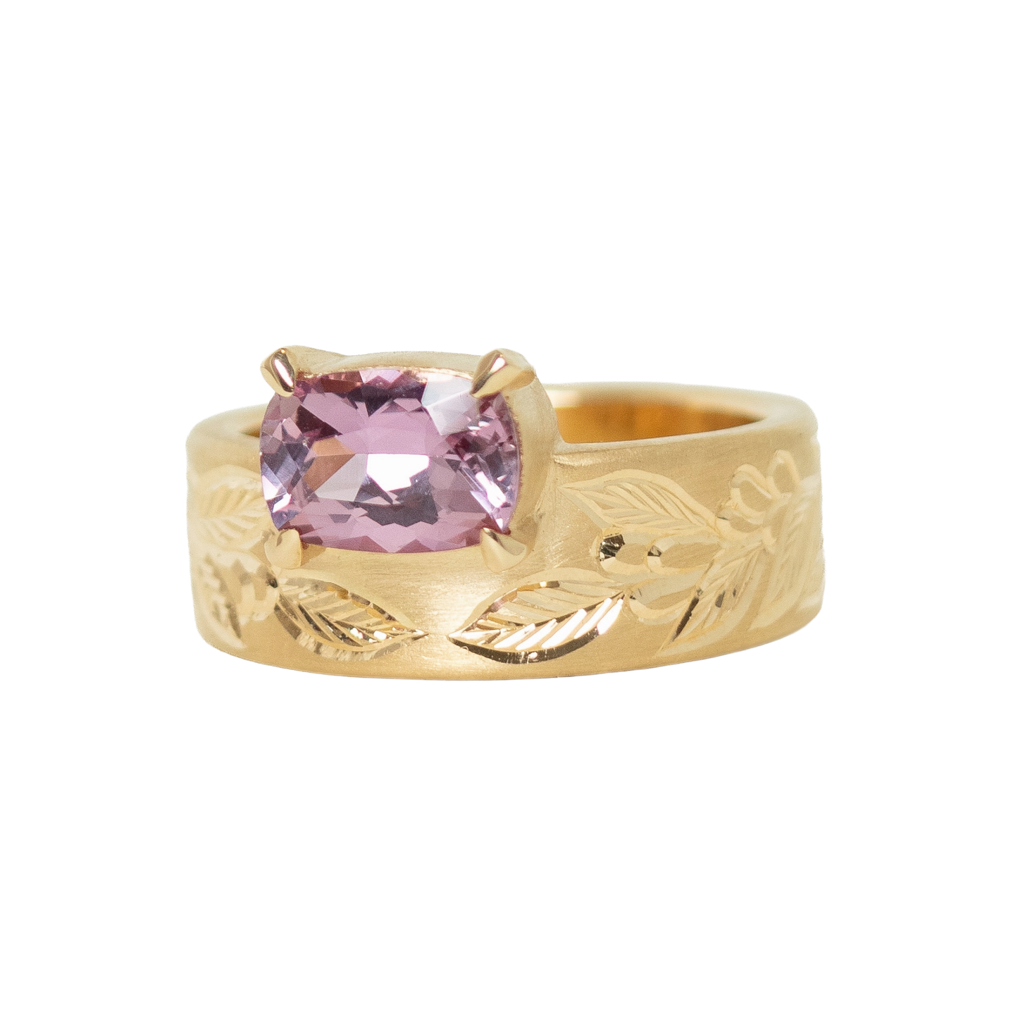A 14k yellow gold ring featuring a cushion-cut pink sapphire in a prong setting, with an hand engraved leaf pattern on the band, shown against a plain white background.