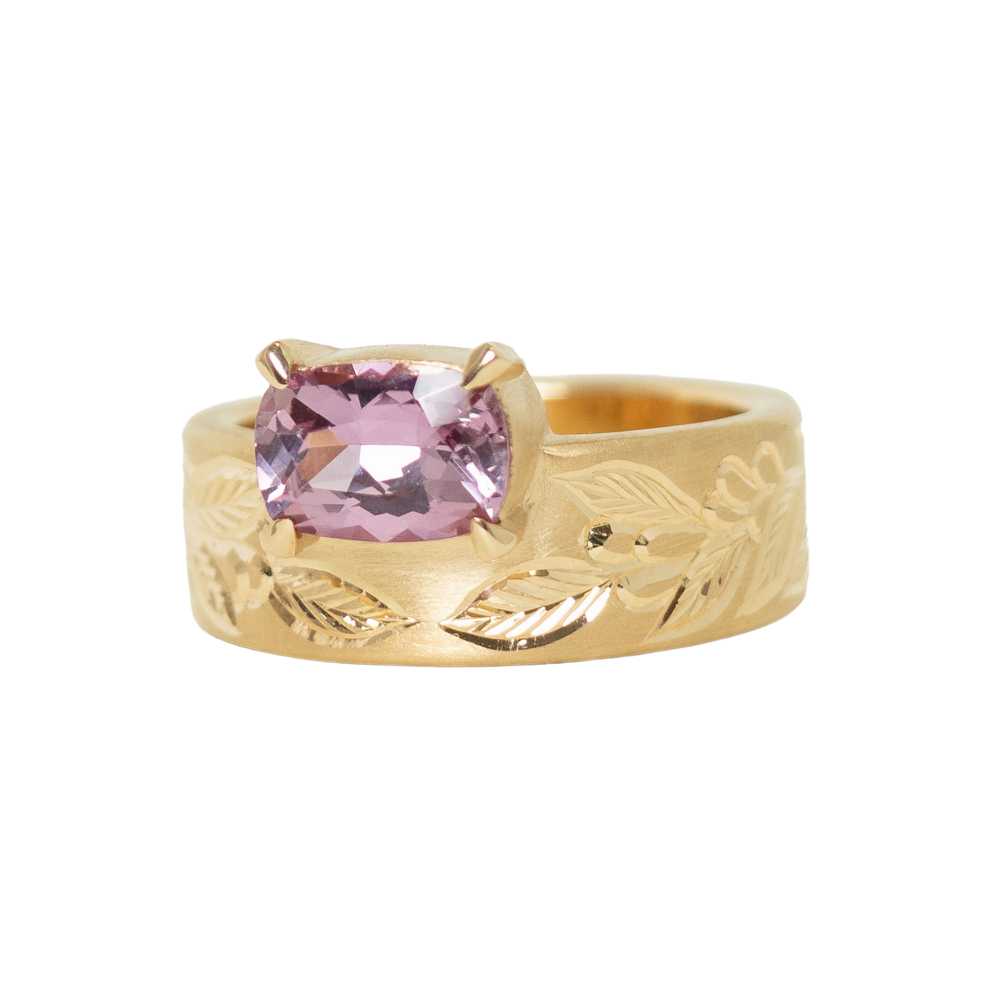 A 14k yellow gold ring featuring a cushion-cut pink sapphire in a prong setting, with an hand engraved leaf pattern on the band, shown against a plain white background.