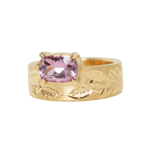 A 14k yellow gold ring featuring a cushion-cut pink sapphire in a prong setting, with an hand engraved leaf pattern on the band, shown against a plain white background.